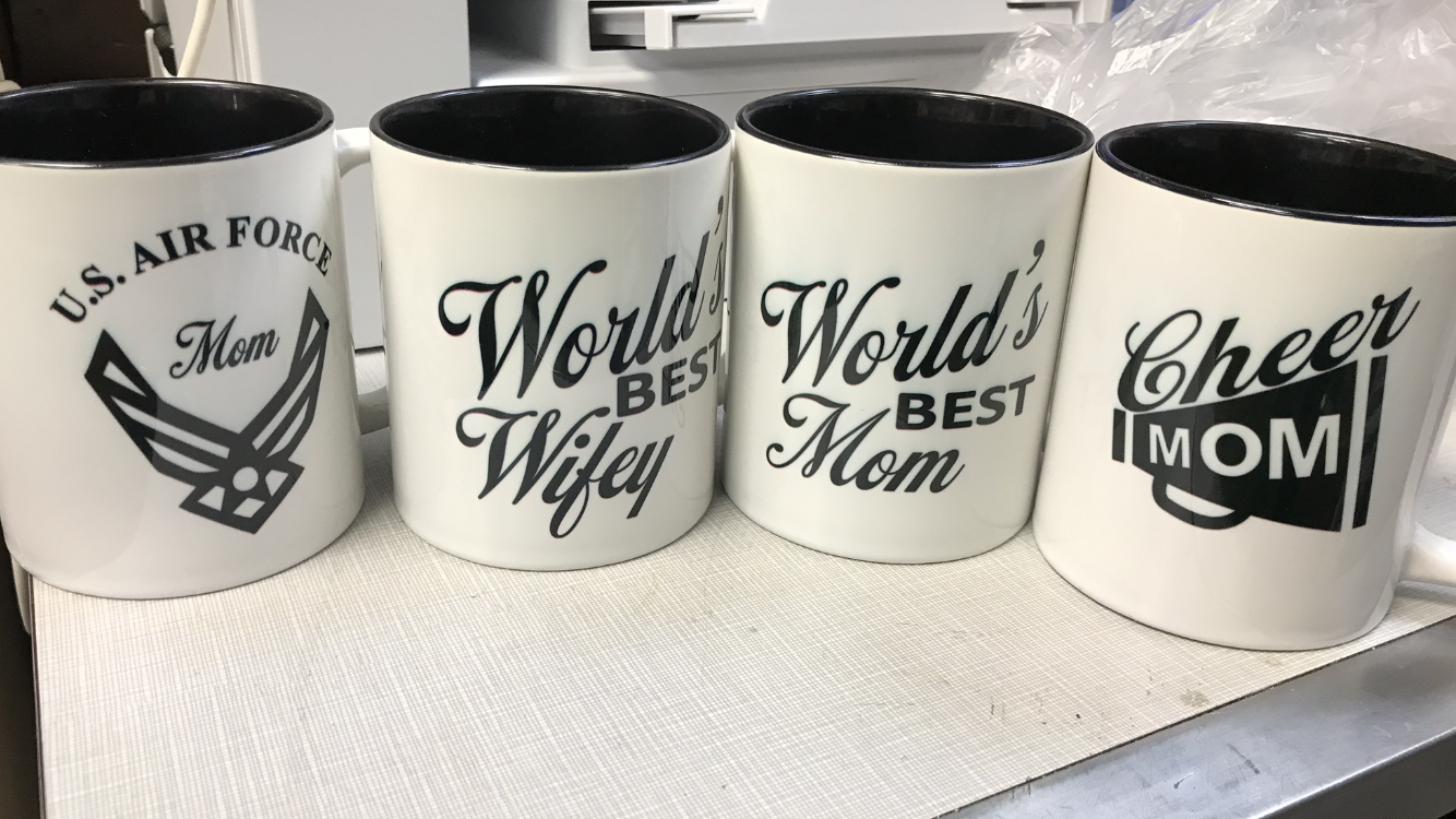 A row of custom engraved mugs reading World's Best Wifey, World's Best Mom, Cheer Mom, and US Air Force Mom